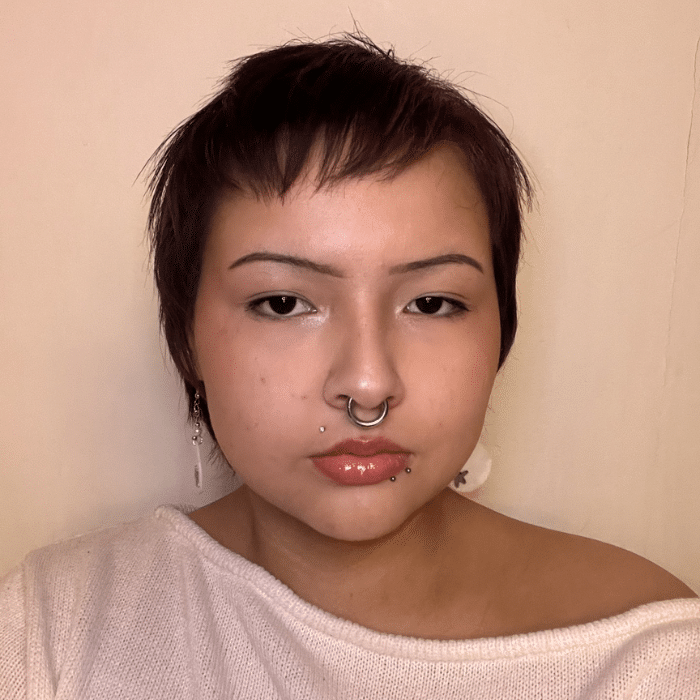 A person with short dark hair and multiple facial piercings looking directly at the camera against a beige wall. She is wearing a white off-the-shoulder top and feather earrings.