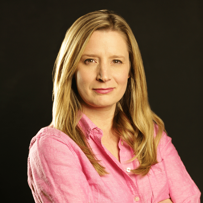 A person with long blonde hair stands with arms crossed, looking at the camera, wearing a bright pink button-up shirt against a dark background.