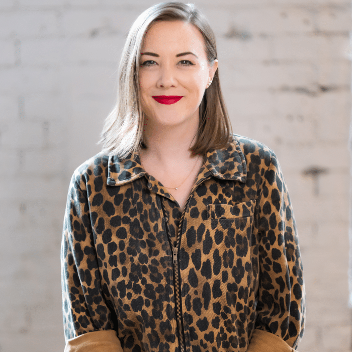 A person with shoulder-length light brown hair smiling at the camera. She is wearing a leopard-print zip-up shirt and bright red lipstick. There is a blurred white brick wall behind her.