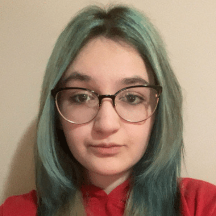 A person with long teal hair and glasses looking at the camera against a neutral wall. She is wearing a red hoodie.