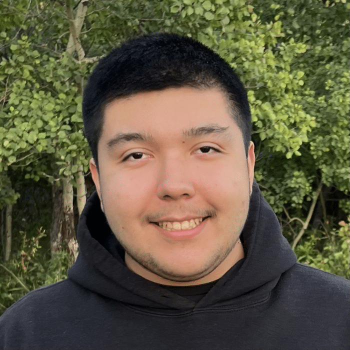 A person with short dark hair and a close-shaved beard smiling in front of green trees outdoors. He is wearing a black hoodie.