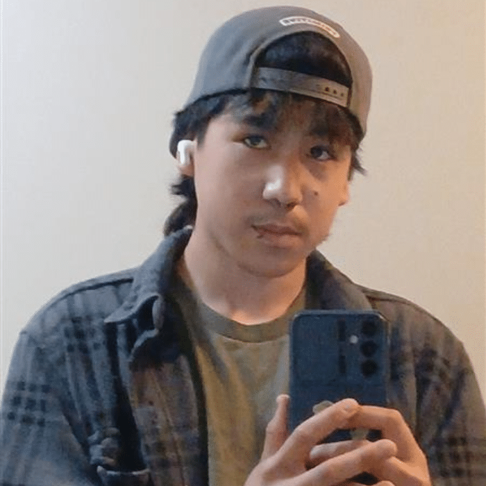 A person with medium-length dark hair and a lip piercing taking a mirror selfie. He is wearing a grey plaid shirt, green T-shirt, and backwards cap, with white earbuds.