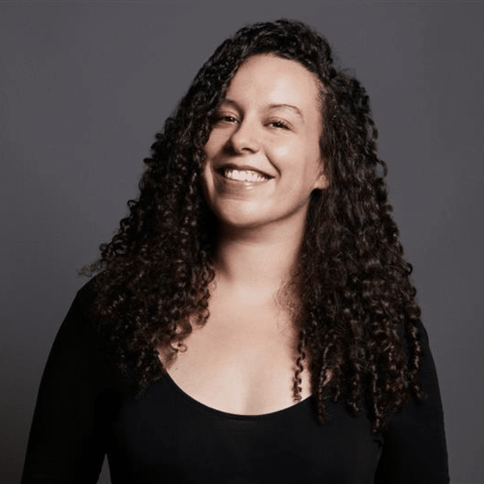 A person with long, dark, curly hair smiles at the camera against a plain grey background. The person is wearing a long-sleeve black shirt.