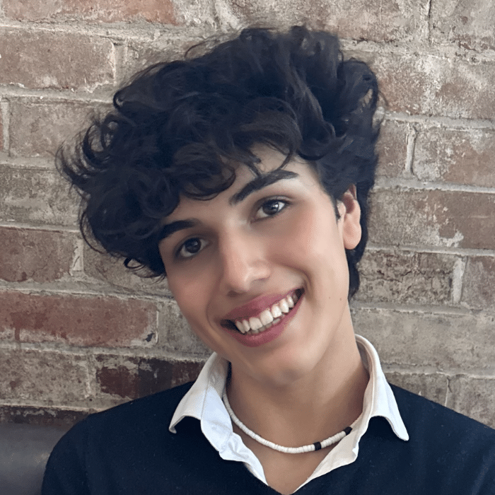 A person with short curly dark hair smiling in front of a brick wall. He is wearing a black sweater over a white collared shirt . They are also wearing a white beaded necklace.