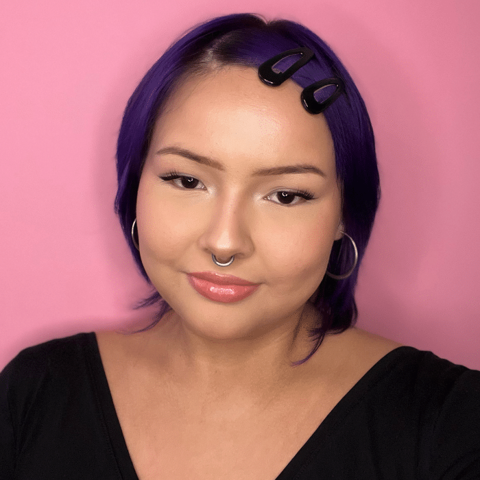 Lindsay Larocque posing against a pink background with purple hair, hoop earrings and a septum ring