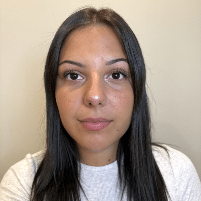 A person with straight dark hair looking directly at the camera against a beige wall. She is wearing a white top.