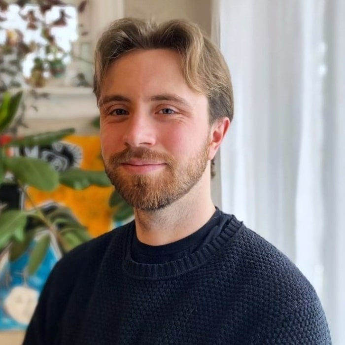 A young man with light brown hair and a short beard smiles gently at the camera. He is wearing a dark knit sweater and standing indoors near a window with soft natural light. Green plants and a colourful abstract artwork are visible in the background.
