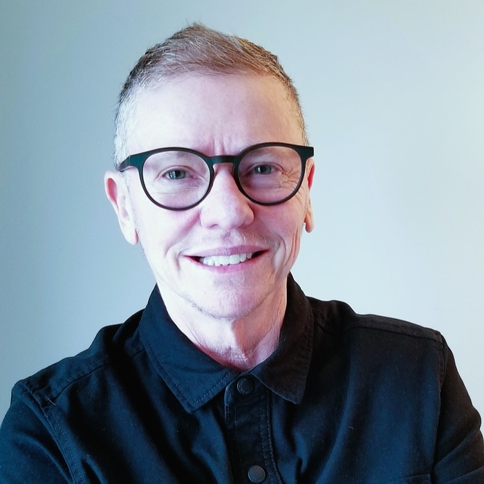 A person with very short, light-coloured hair and round black glasses smiles at the camera. They are wearing a black button-up shirt and are photographed against a plain, light grey background with soft, even lighting.