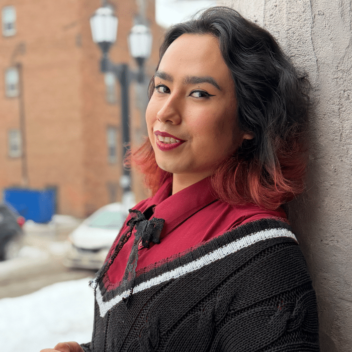 Person with dark hair leaning against a wall outdoors, smiling slightly.