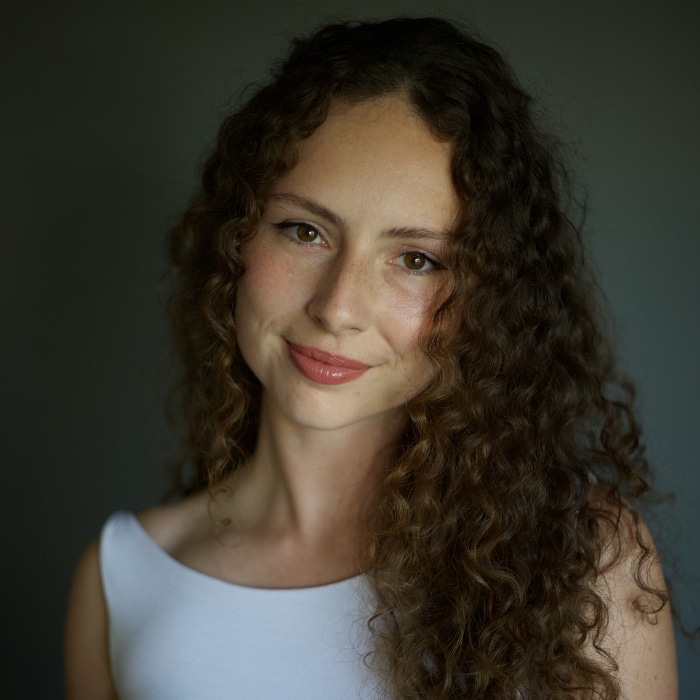 A young woman with long, dark brown curly hair looks directly at the camera with a soft smile. She is wearing a white top and minimal makeup. The background is dark and softly blurred, creating a studio-style portrait.