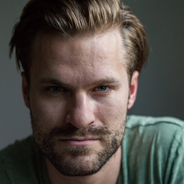 A man with short brown hair and light stubble looks directly into the camera with a serious expression. He is wearing a muted green shirt. The background is softly lit and neutral, with a shallow depth of field.