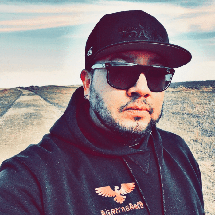 Person wearing a black sweatshirt, sunglasses and a cap outdoors on a road.