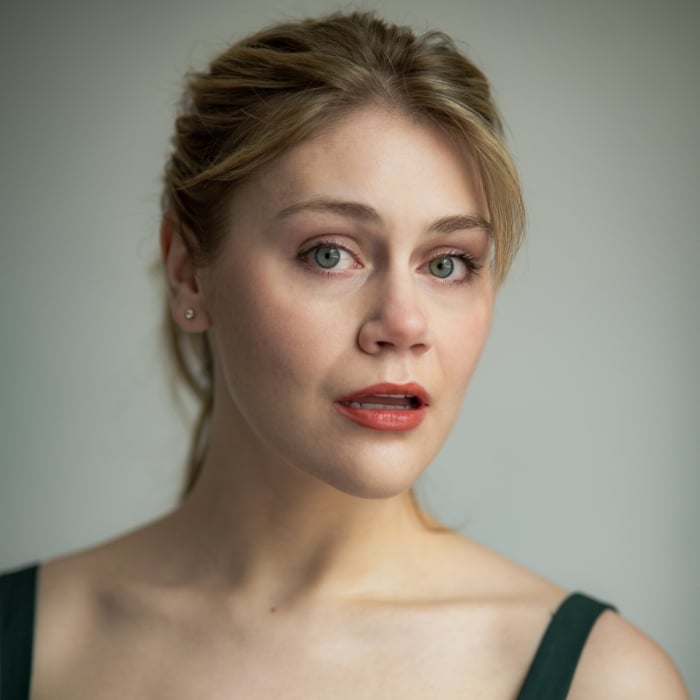 A young woman with blonde hair pulled back looks at the camera with a slightly surprised or attentive expression. She is wearing a dark green sleeveless top and small stud earrings. The background is softly lit and neutral.