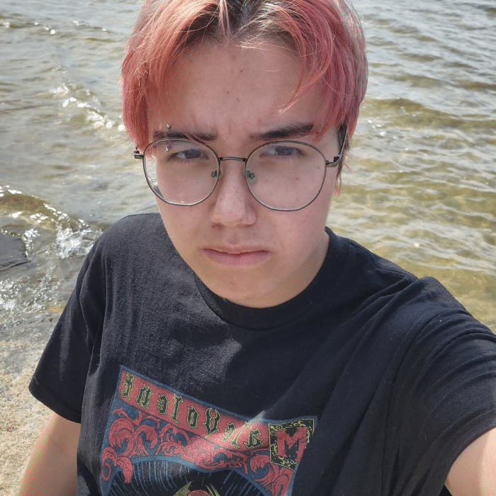 Person with short pink hair and glasses standing in water, looking serious.