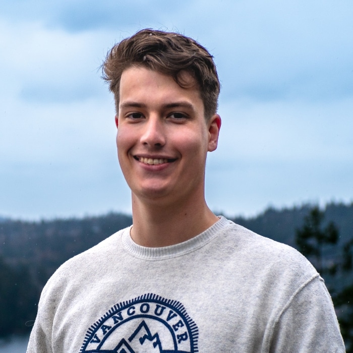 A young person with short brown hair smiles at the camera while standing outdoors. They are wearing a light grey sweatshirt with a Vancouver graphic. A cloudy sky and a blurred natural landscape appear in the background.