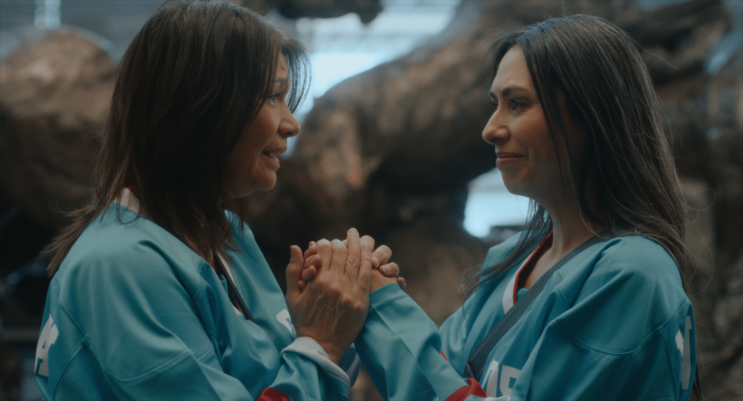 Two people in teal hockey jerseys stand face to face indoors, smiling and holding hands in an emotional moment of connection and support. The background is softly blurred with natural rock formations visible behind them.