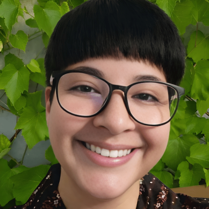 Chey Wright smiling outdoors in front of green leaves wearing glasses and a patterned shirt