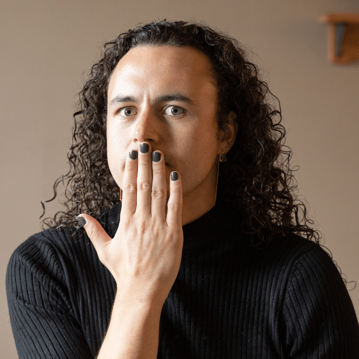 Everett Sokol posing indoors with hand covering mouth, wearing a black sweater and long earring