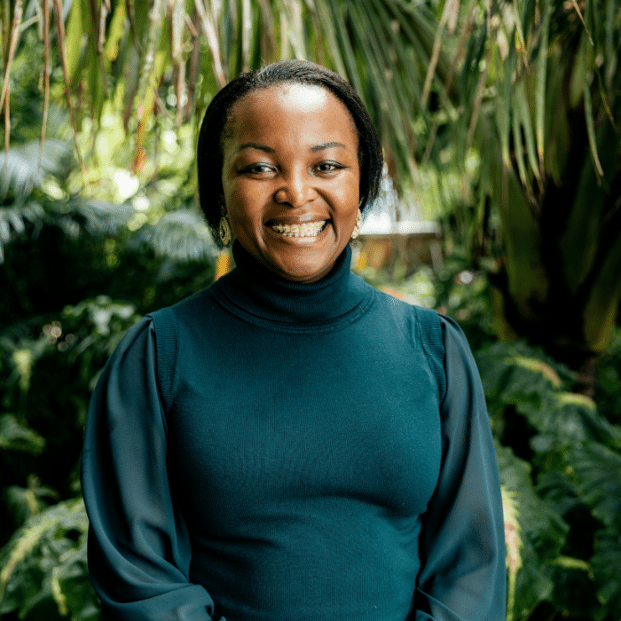 Portrait of a smiling person standing outdoors in a lush garden setting, wearing a dark green high-neck top with sheer sleeves and small earrings. The background is filled with tropical greenery and soft natural light.
