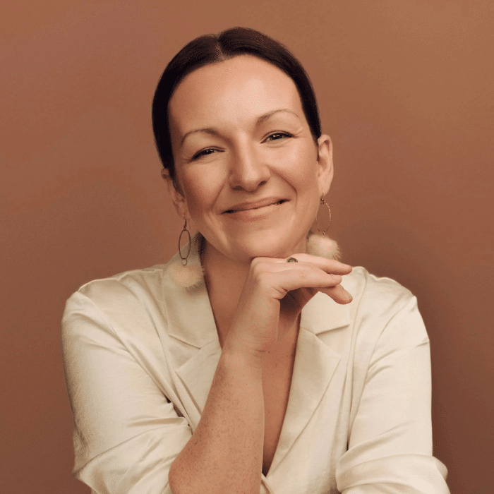 A smiling woman with dark hair pulled back poses against a warm brown background, resting her chin on one hand and wearing a cream-coloured top with dangling earrings.