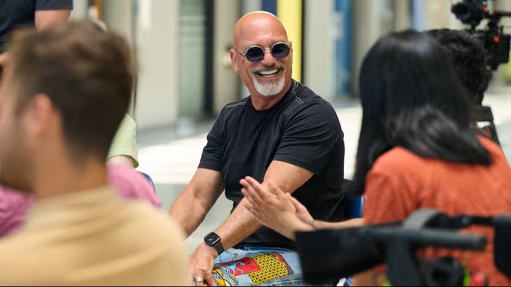 A smiling man in round sunglasses participates in a group conversation while seated among other attendees in a bright indoor space.